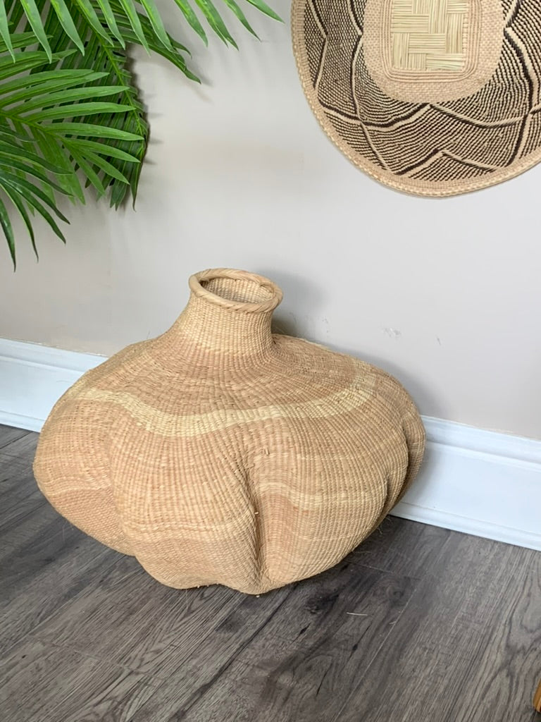 A large, hand-woven natural fiber vase with a bulbous, rounded shape and a short neck, styled on a dark wood floor against a beige wall with a green plant in the background.