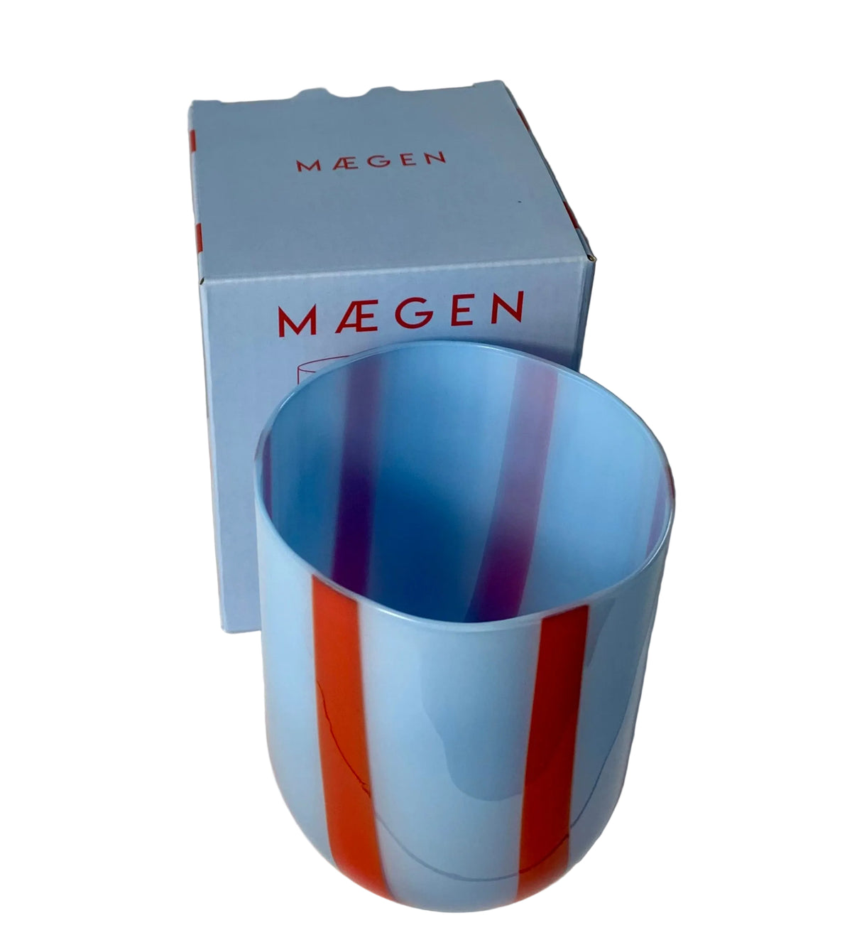 A light blue glass tumbler with vertical red and purple stripes, shown in front of its light blue MÃEGEN box on a white round surface against a white fabric backdrop.