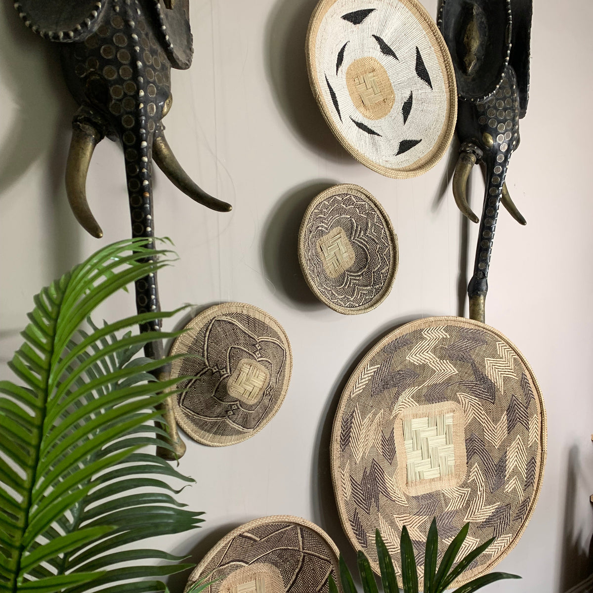 A styled interior wall display featuring multiple circular woven and carved decorative pieces in earthy beige, brown, and black tones, alongside two dark elephant wall sculptures and green palm leaves in the foreground.