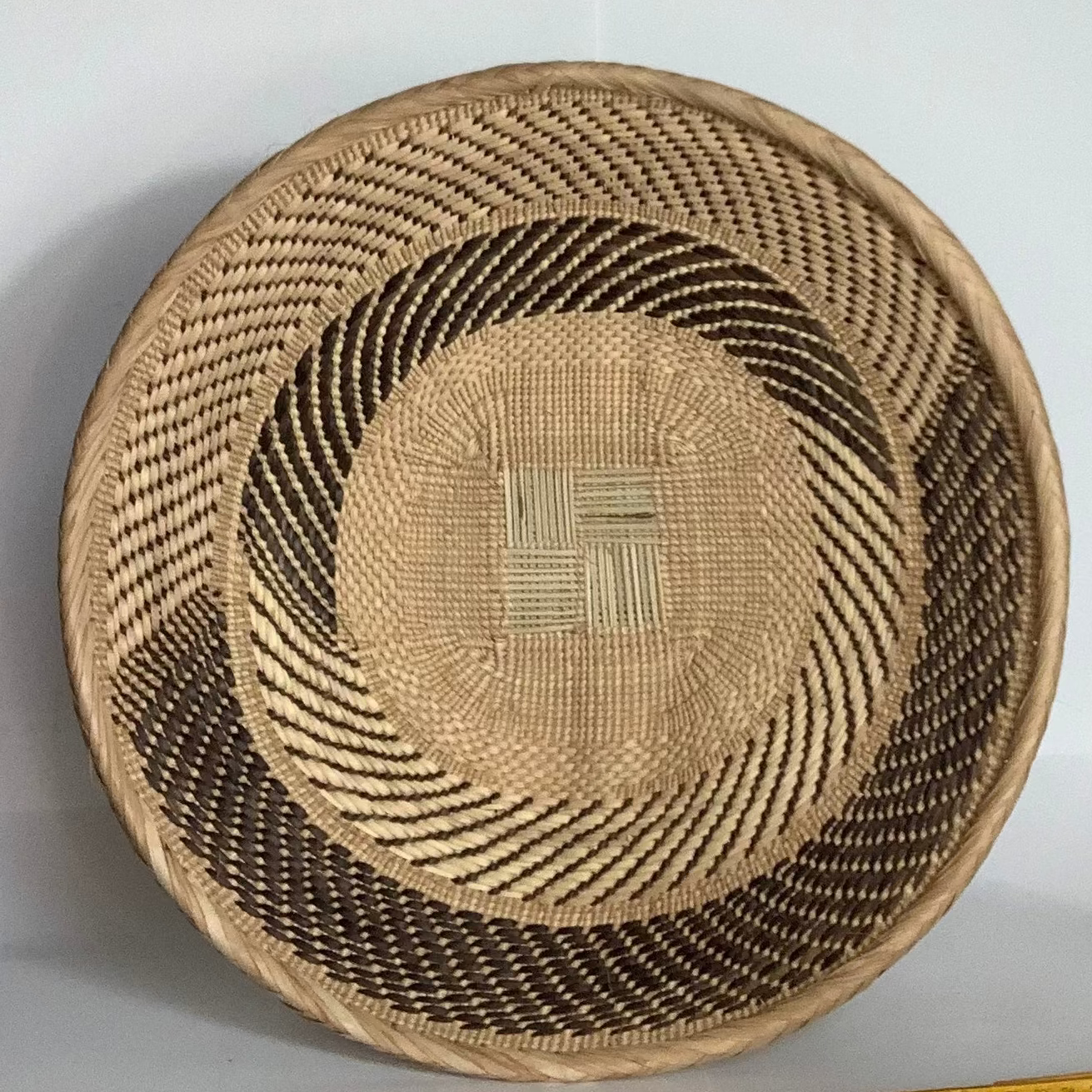 A round woven basket with a natural tan base and dark brown concentric patterned bands, shown against a white background with a yellow measuring tape ruler at the bottom.