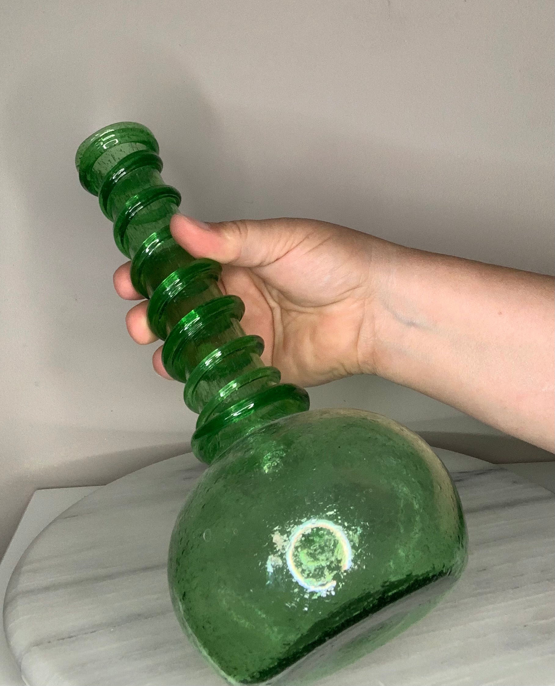 A hand holding a green glass bottle with a ribbed, spiral-shaped neck and a rounded base, set against a neutral indoor background.