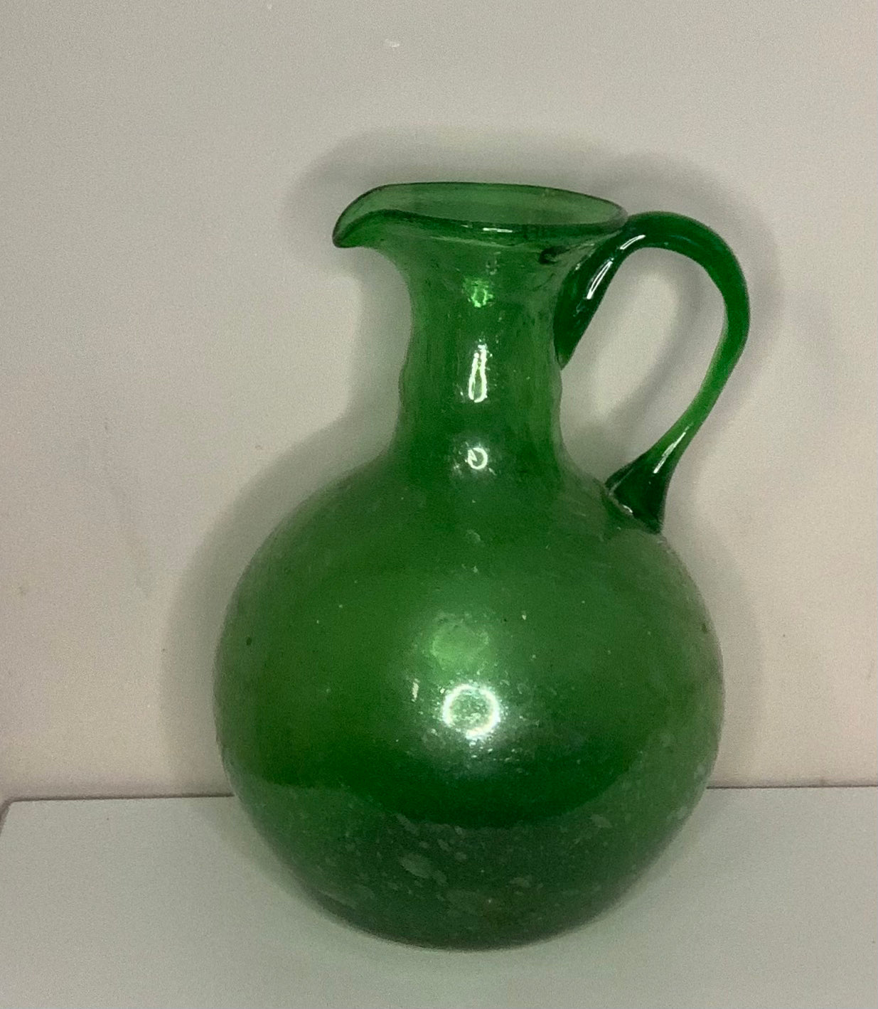 A vintage green glass pitcher with a rounded body, short neck, and curved handle, shown against a plain off-white background.