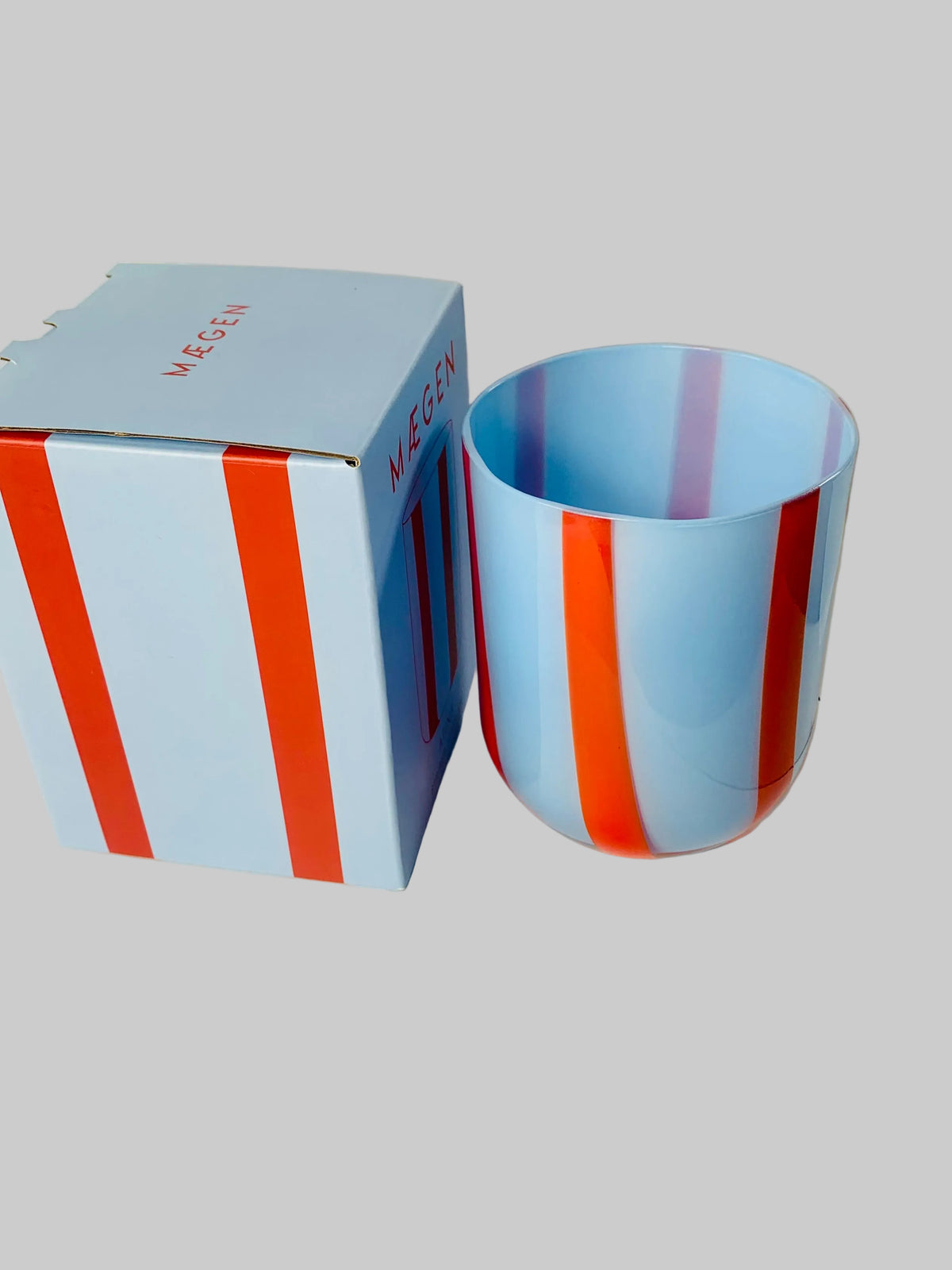 A light blue MEGEN glass cup with vertical red and purple stripes, shown next to its matching light blue and red striped box on a white circular display stand.