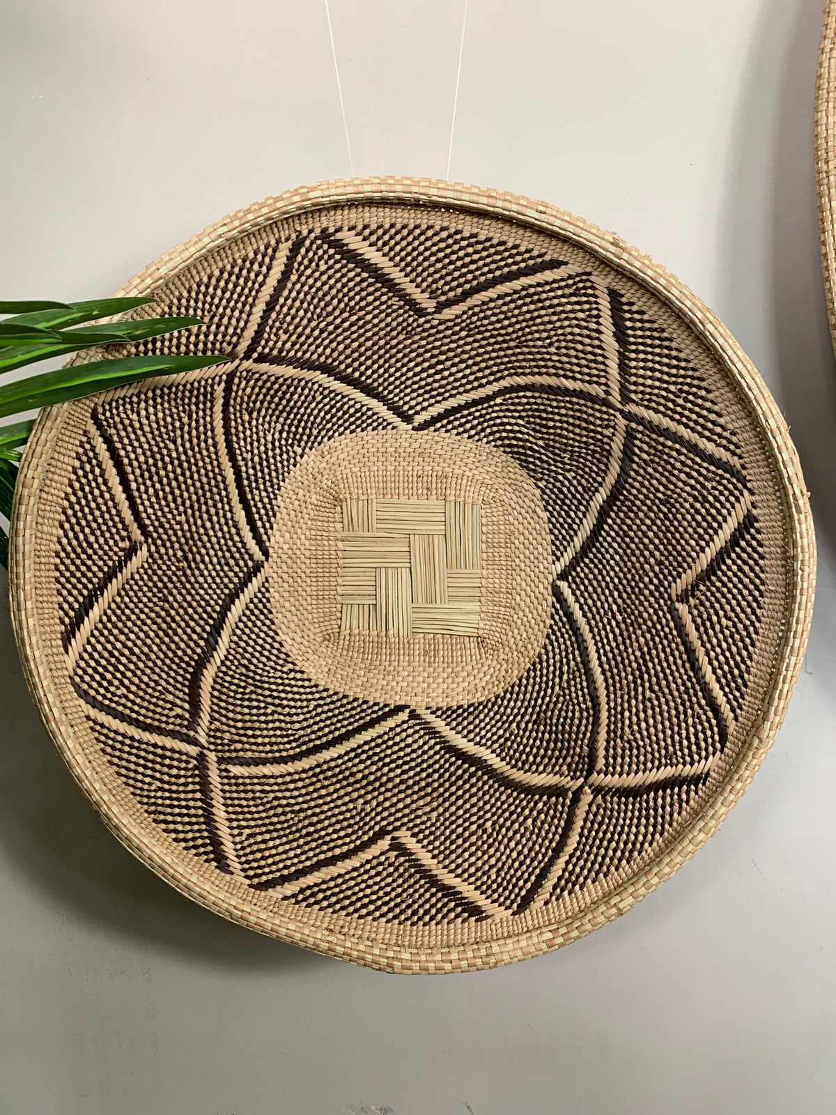 A round woven basket made from natural straw and dark brown thread, featuring a geometric pattern and a central woven medallion, hanging against a light gray wall.