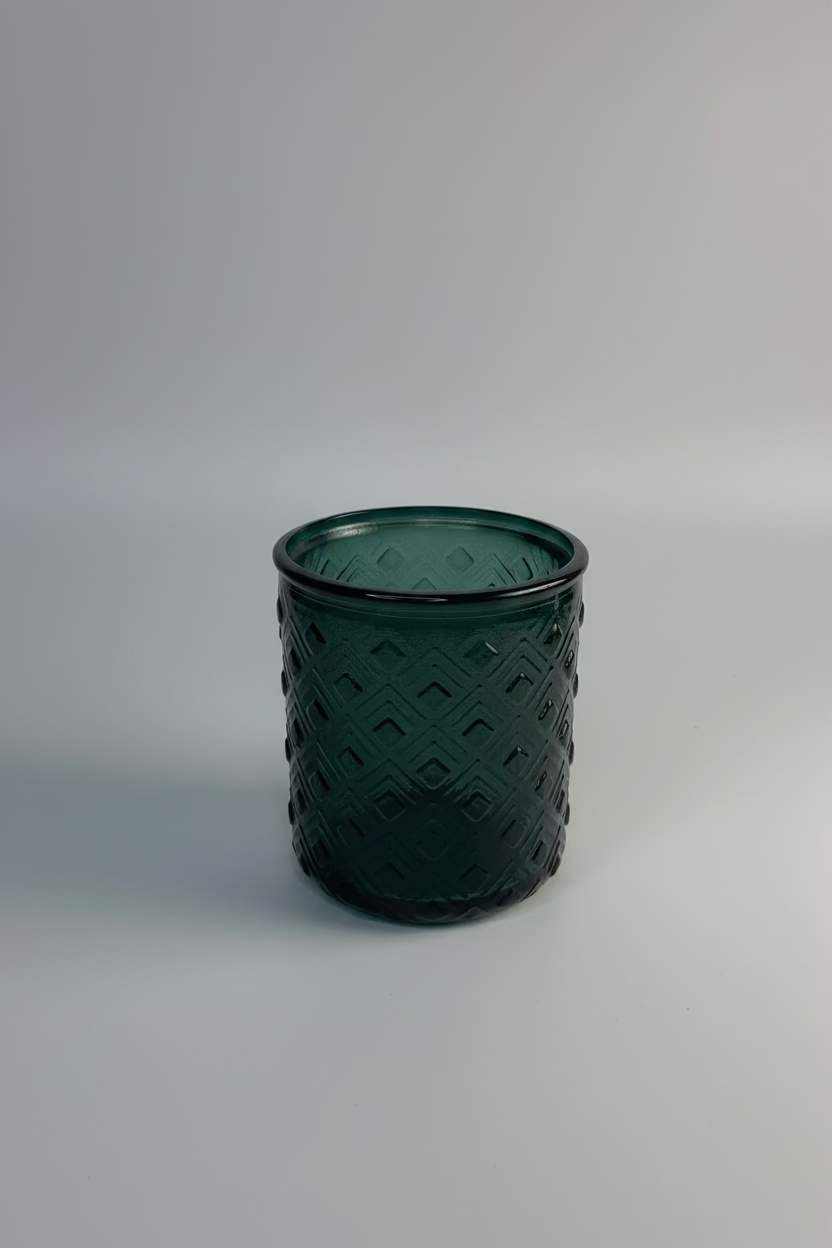 A dark green textured glass tumbler with a diamond pattern, displayed on a white circular riser against a draped white fabric background.