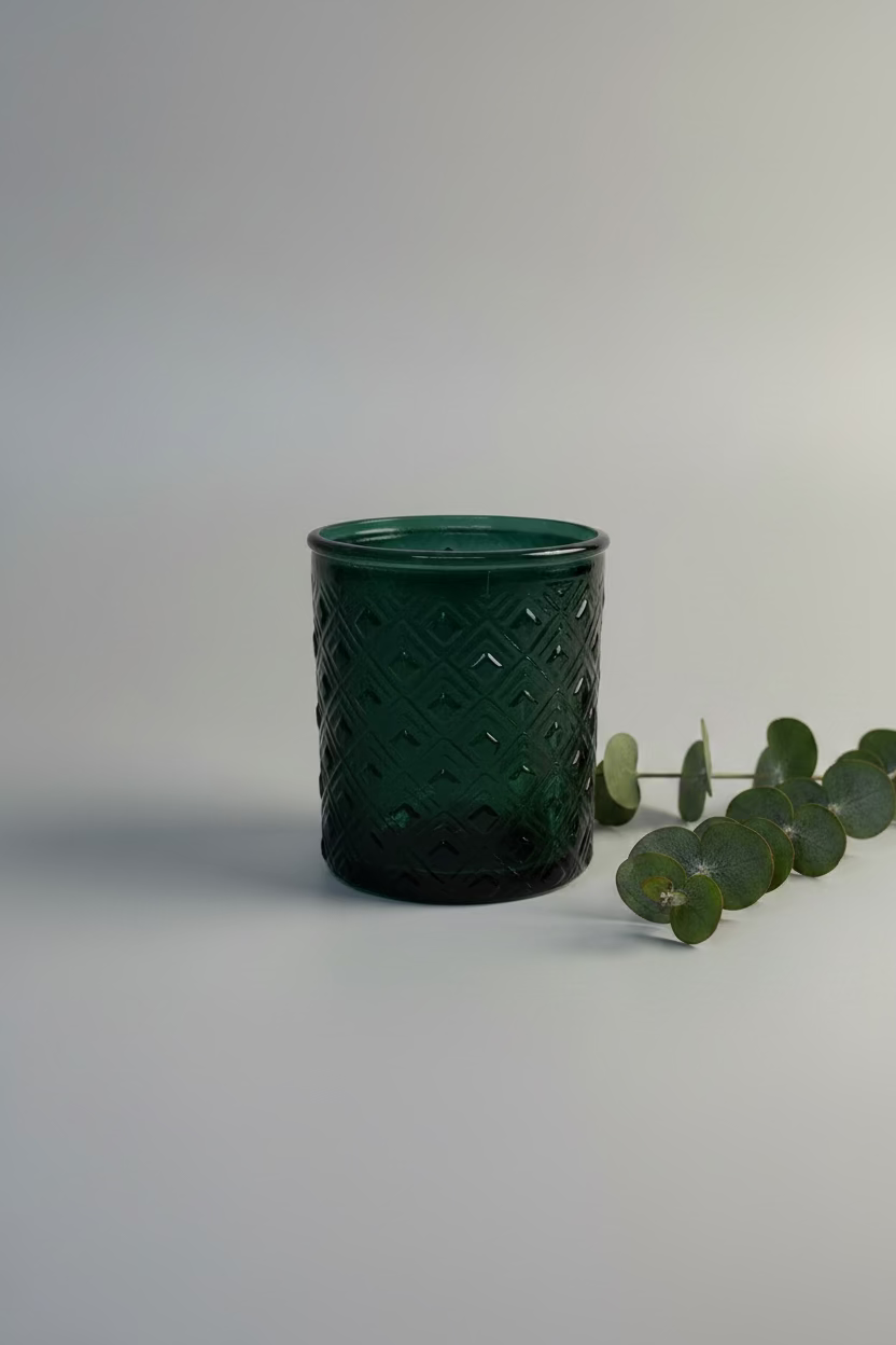 A dark green textured glass tumbler with a diamond-patterned interior, displayed on a white round pedestal with eucalyptus leaves 