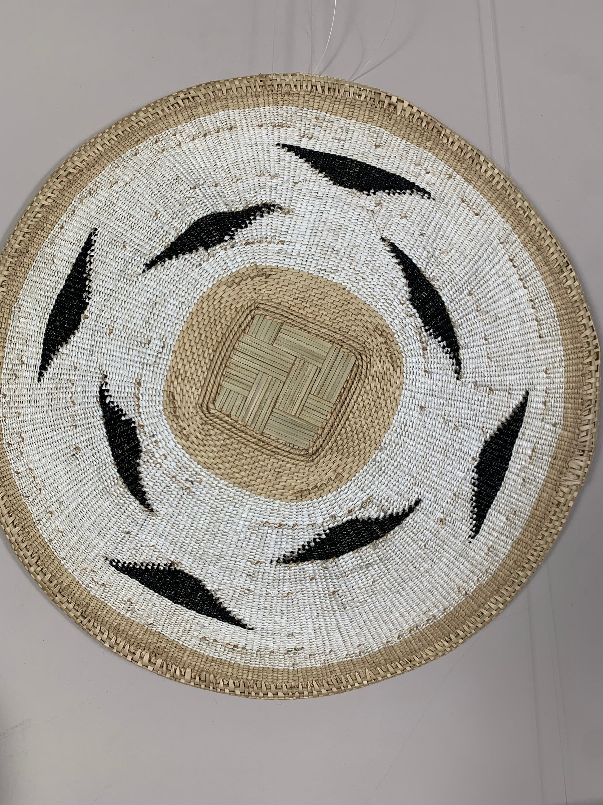 A round woven basket or decorative mat with a natural beige center, white woven sections, and black leaf-like accents, photographed against a plain light background.