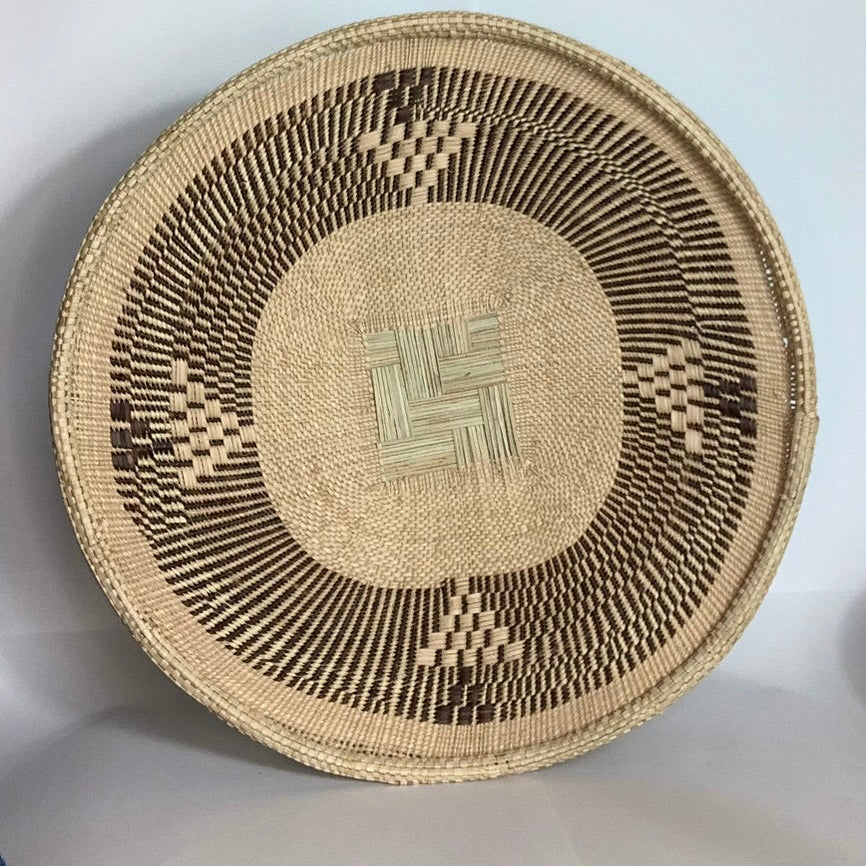 A round woven basket made from natural tan palm, featuring a geometric pattern in dark brown and light beige, photographed against a plain light gray background.