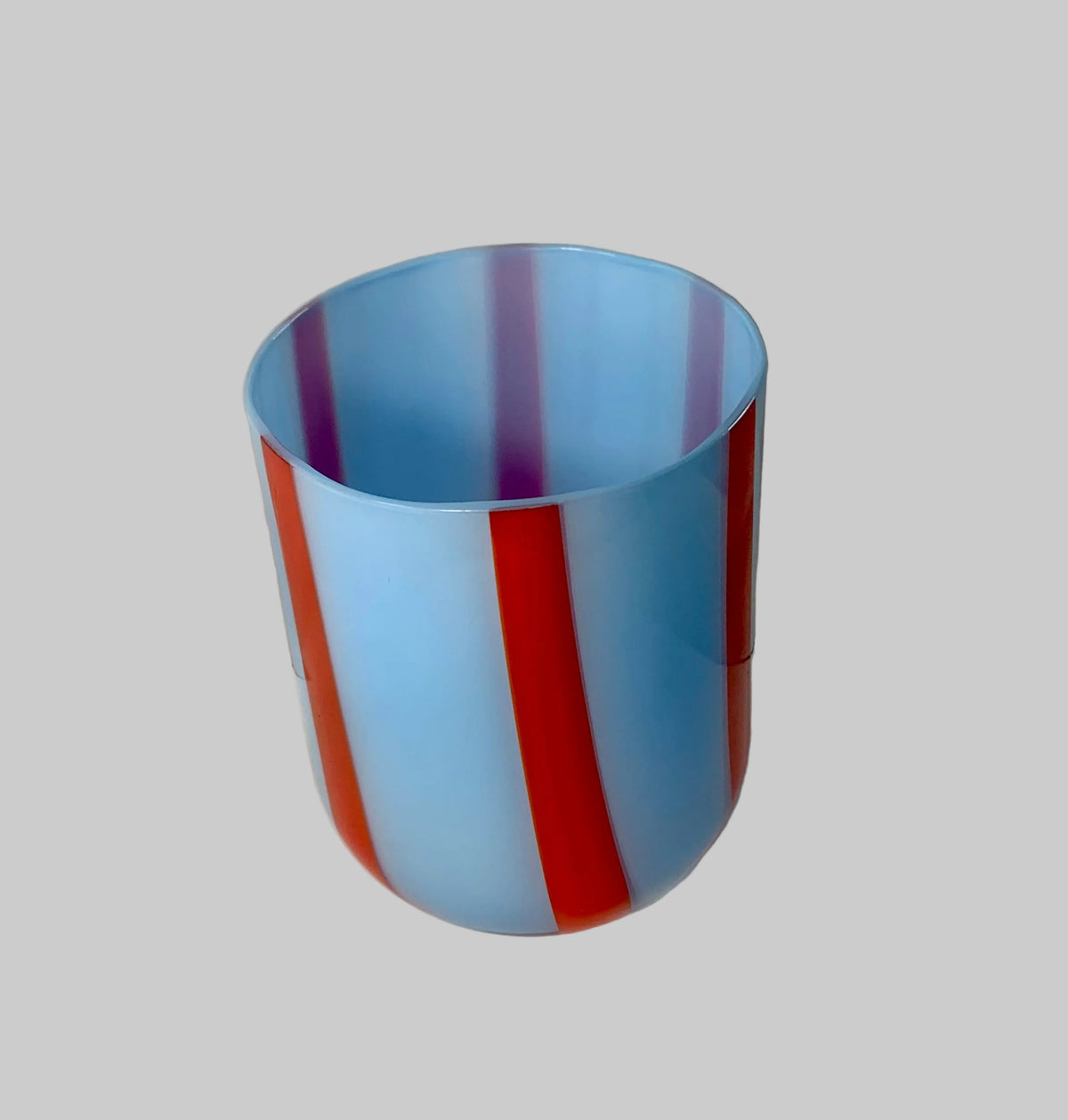 A translucent cylindrical cup or tumbler with vertical stripes in light blue, red, and purple, photographed on a white round surface against a wrinkled white backdrop.
