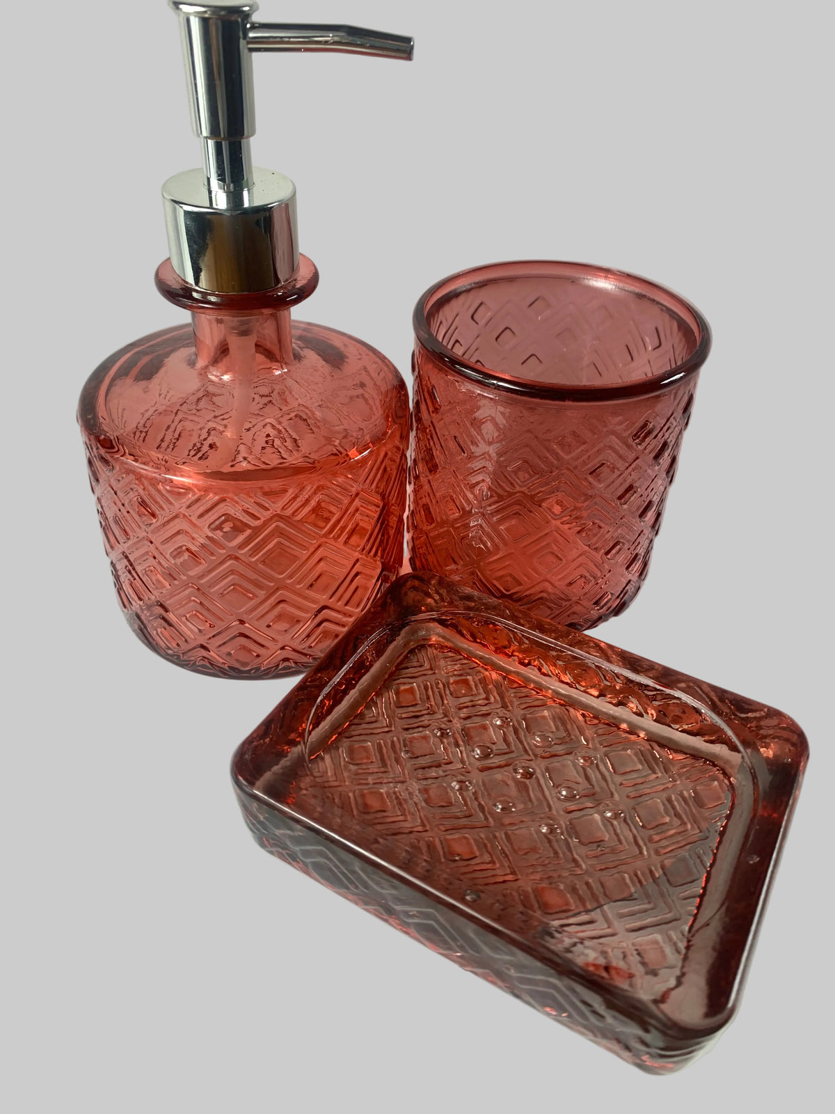 A matching set of three pink patterned glass bathroom accessories, including a soap dispenser with a chrome pump, a tumbler, and a rectangular soap dish
